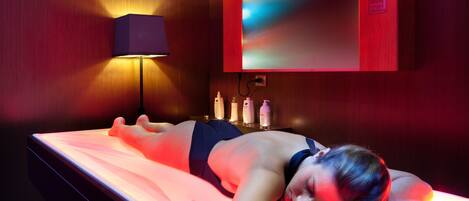 A person lying on a massage table in a room with a wooden wall, a massage lamp, and a digital device on the table.