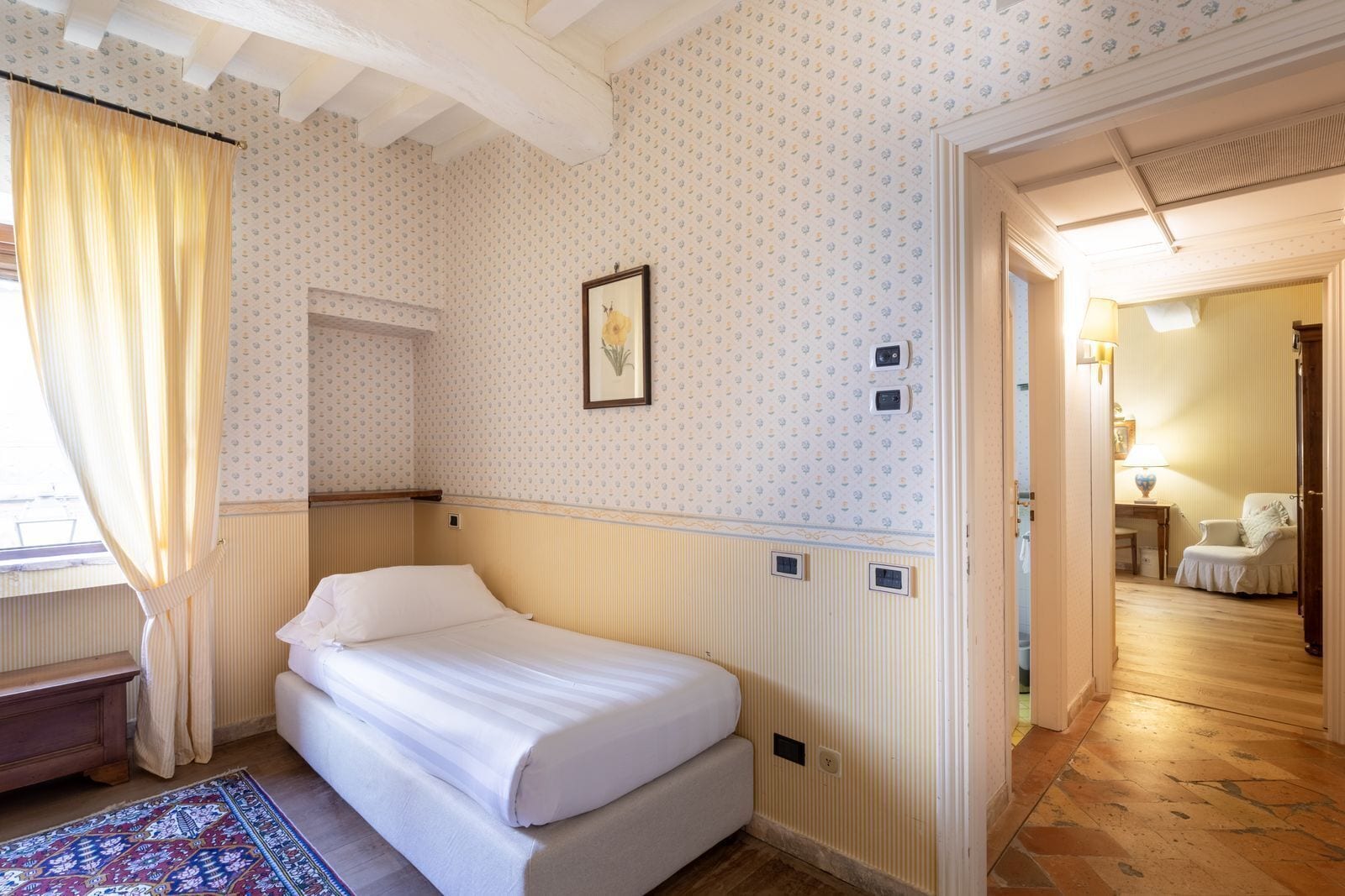 Una habitación de hotel con cama, ventana con cortinas, papel tapiz estampado, un cuadro en la pared y suelo de madera.