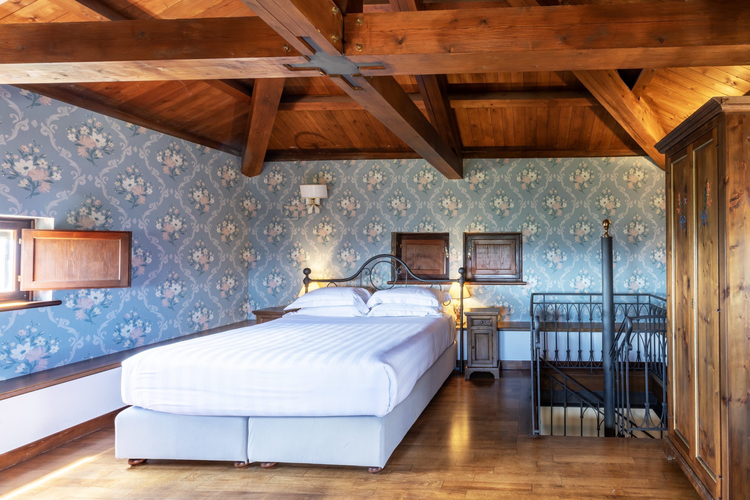 Ein Schlafzimmer mit einem großen Bett, Holzbalkendecke, Blumenpapier und einem Nachttisch. Eine Lampe und eine Treppe sind ebenfalls vorhanden.