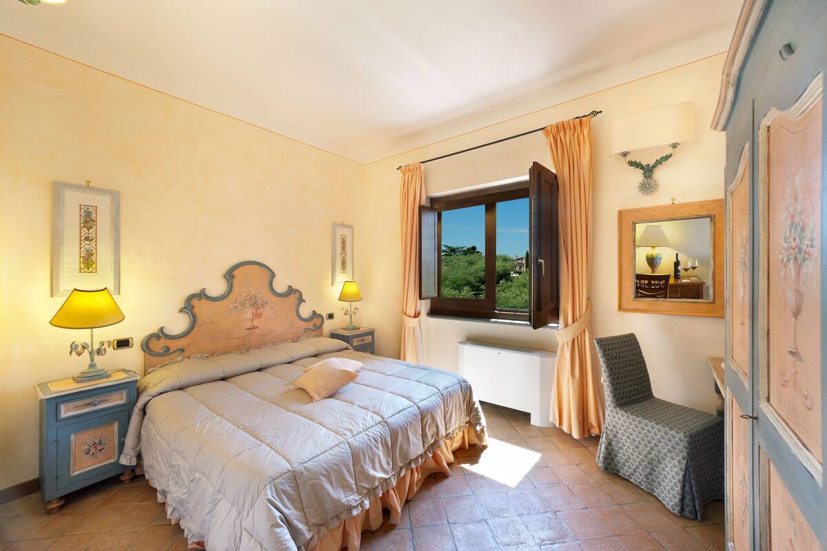 A bedroom with a bed, nightstands, a chair, a window with curtains, and wall-mounted lamps.