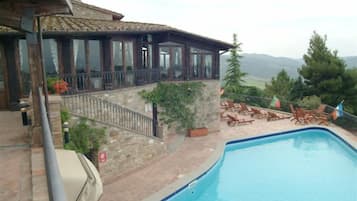 Zona de piscina con un edificio de piedra, un balcón con barandillas de vidrio y vistas a las montañas.