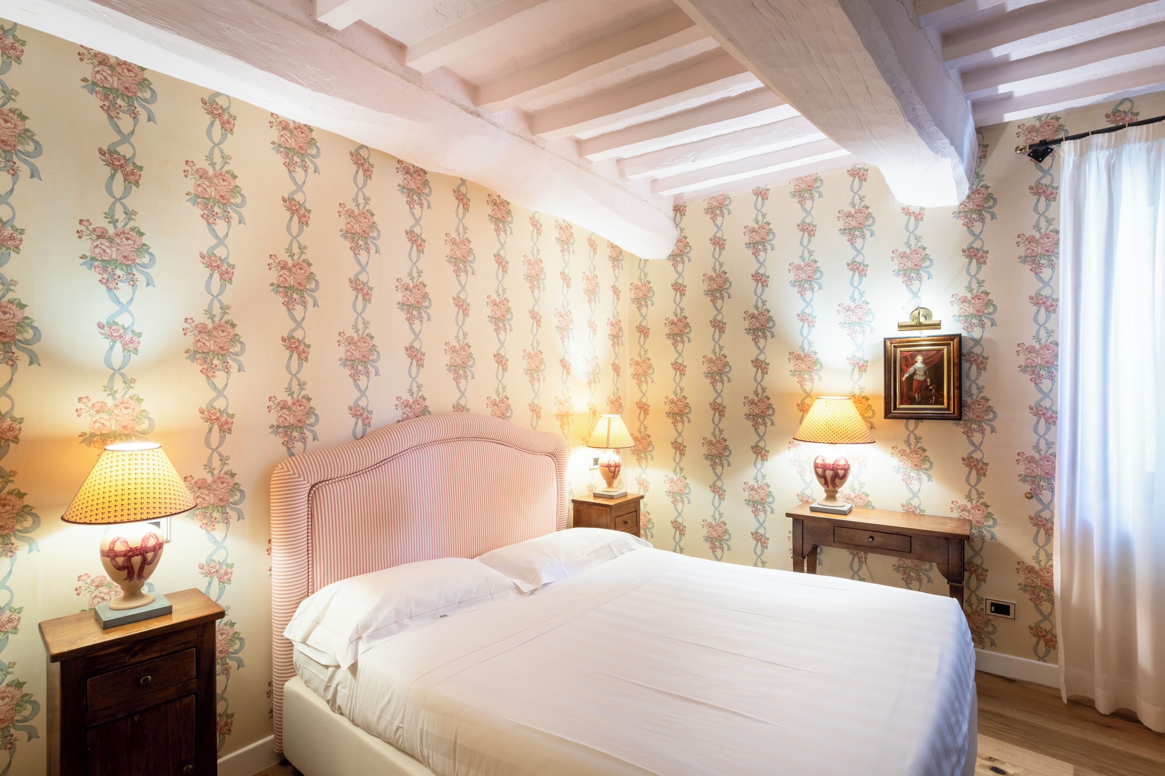 Une chambre à coucher avec un papier peint à motifs floraux, un lit recouvert de draps blancs, deux tables de chevet avec des lampes et un plafond avec des poutres apparentes.