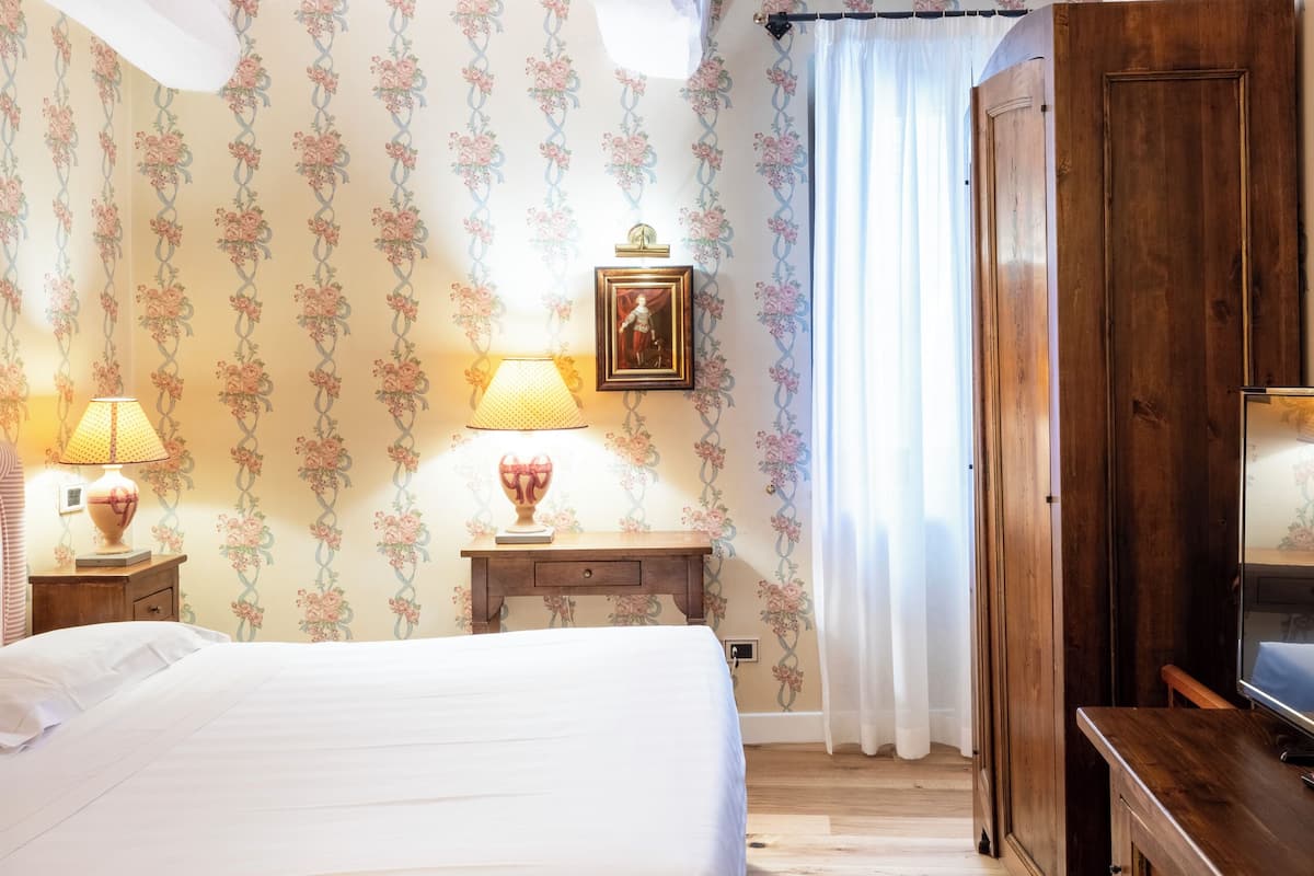A bedroom with a bed, two bedside tables with lamps, a wooden wardrobe, and a floral wallpaper pattern.