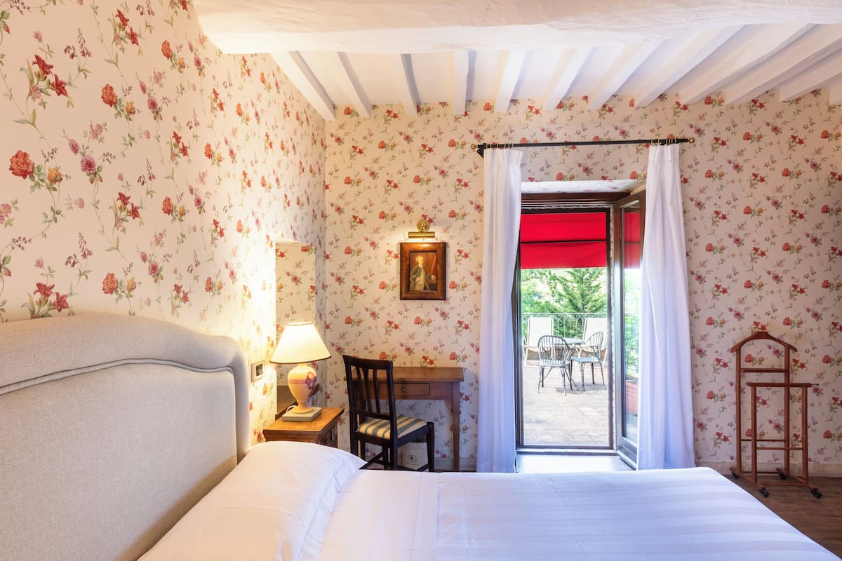 A bedroom with floral wallpaper, a bed, a desk with a chair, and a window with a red curtain.