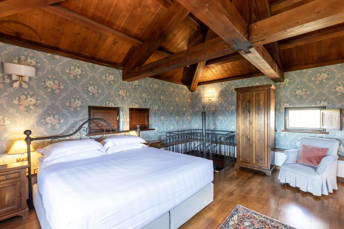 A bedroom with a large bed, wooden ceiling beams, floral wallpaper, a nightstand, a chair, and a cabinet.