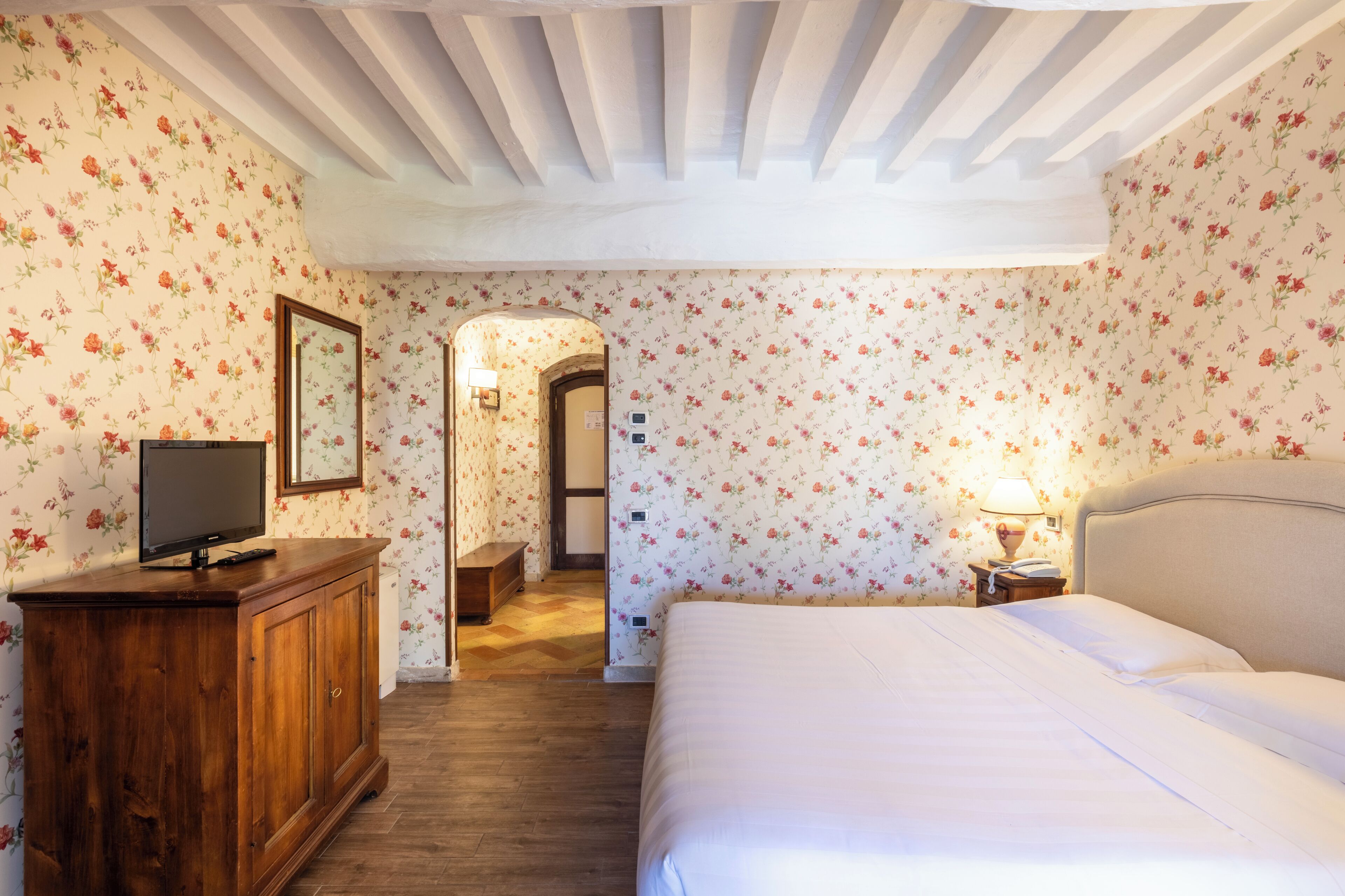 Un dormitorio con cómoda de madera, televisión, cama con ropa blanca, mesita de noche con lámpara y papel tapiz floral.