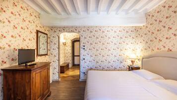 Un dormitorio con cómoda de madera, televisión, cama con ropa blanca, mesita de noche con lámpara y papel tapiz floral.