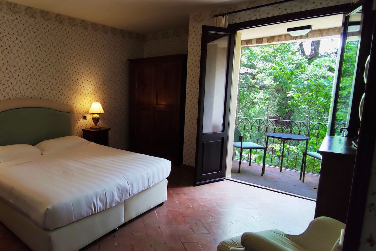 A bedroom with a bed, two bedside lamps, a chair, and a view of a balcony with outdoor furniture.