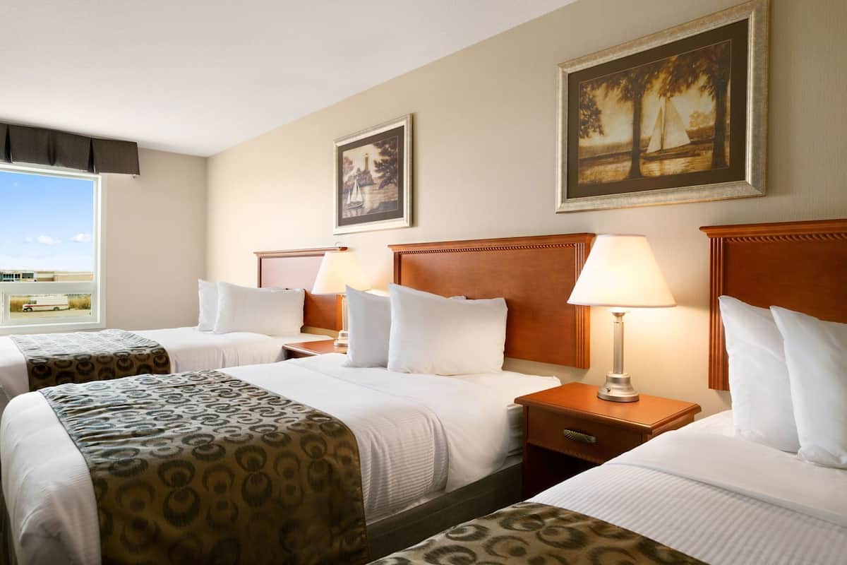 Quarto de hotel com três camas, uma janela grande e duas pinturas nas paredes.