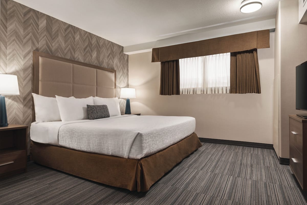 A hotel room with a large bed, a headboard, a nightstand, a lamp, a window with curtains, and a television on a cabinet.