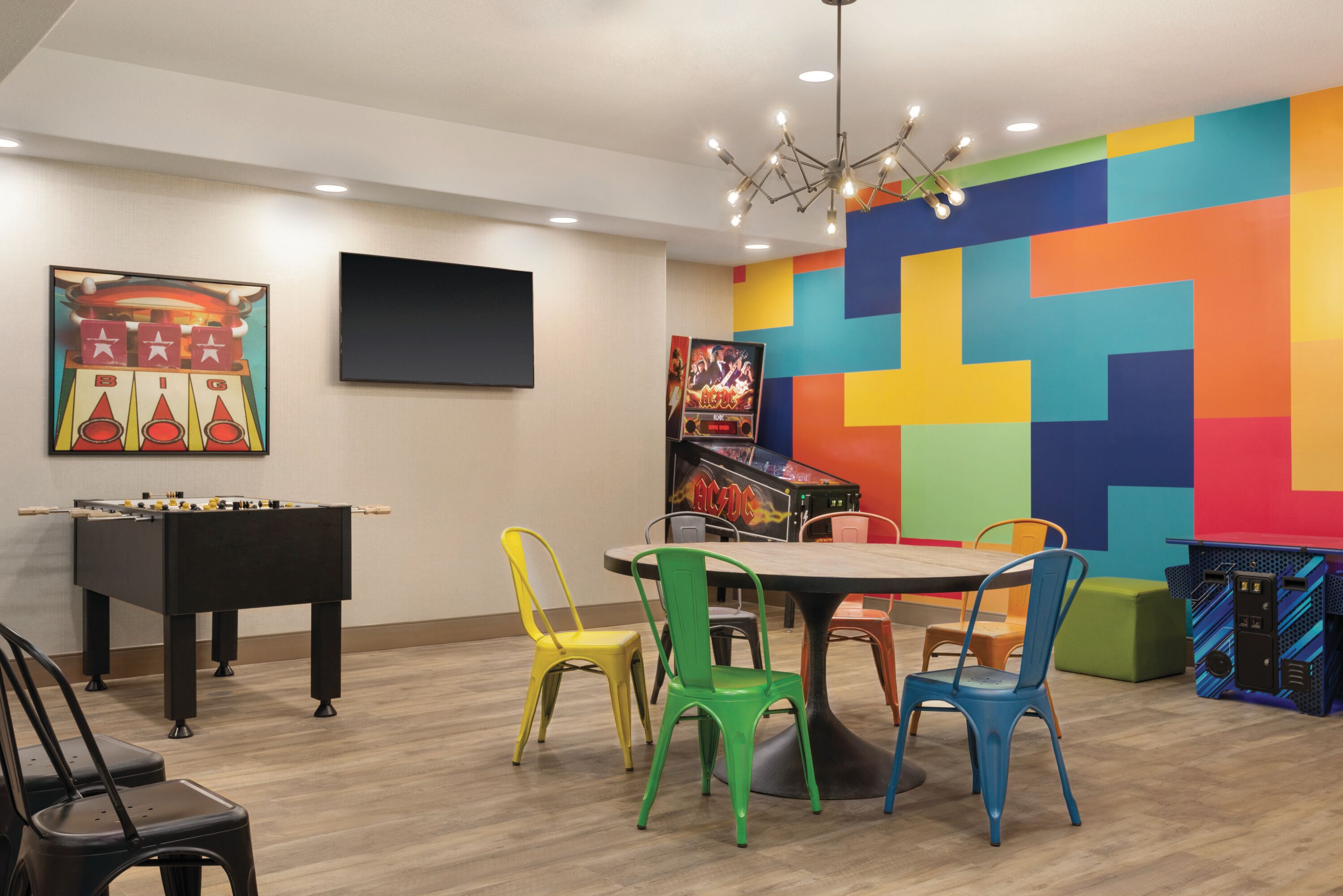 A room with a foosball table, colorful chairs, a table, and a wall with a colorful geometric pattern.
