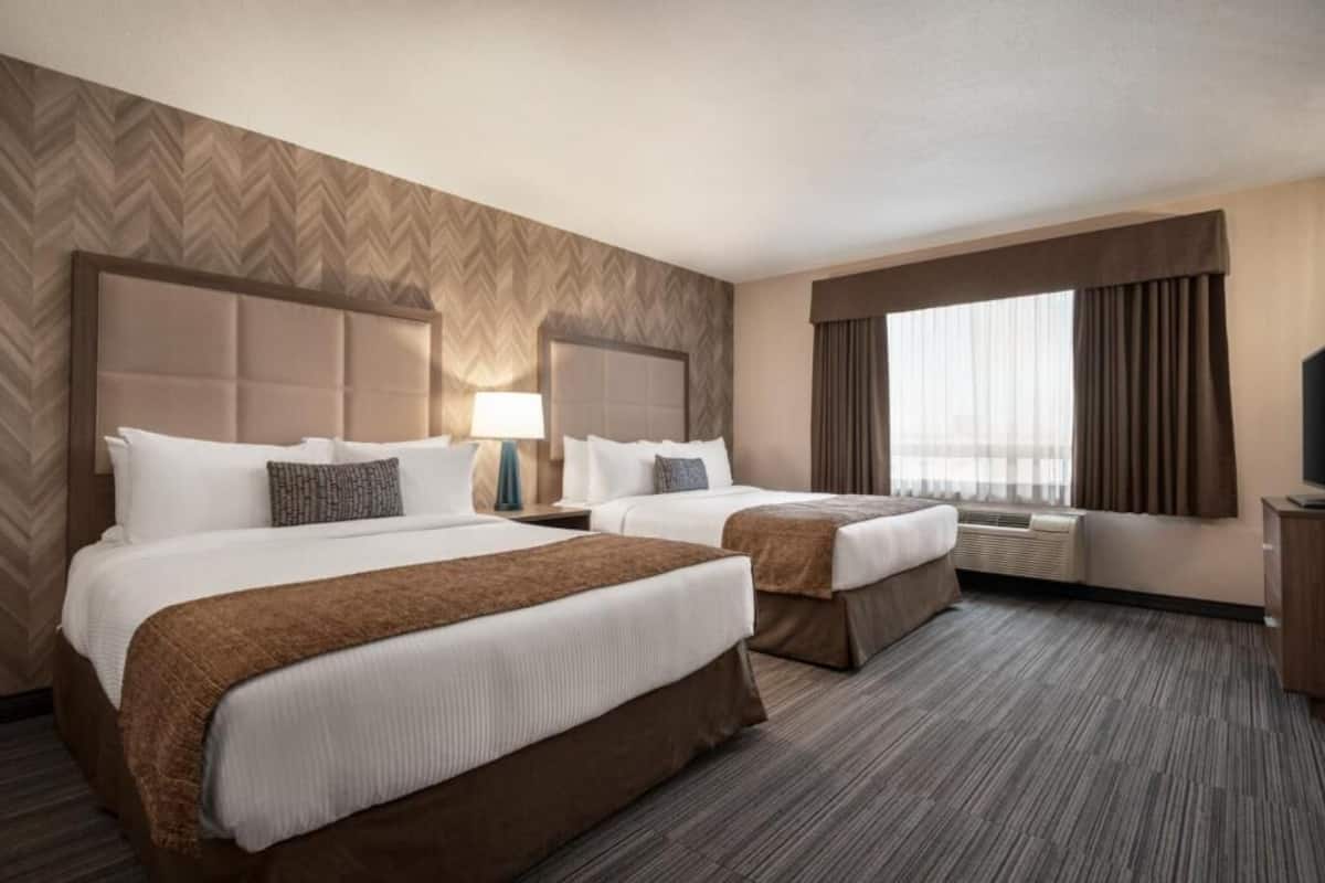 A hotel room with two beds, a large window with curtains, an air conditioning unit, and a television on a wooden cabinet.
