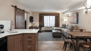 Ein modernes Hotelzimmer mit Küchenzeile, einem Wohnbereich mit Sofa und Esstisch sowie einem großen Fenster mit Vorhängen.