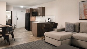 Eine moderne Küche mit dunklen Holzschränken, einem schwarzen Kühlschrank und einer weißen Arbeitsplatte. Ein Essbereich mit Holztisch und -stühlen sowie ein beiges Sofa mit einem gemusterten Kissen.