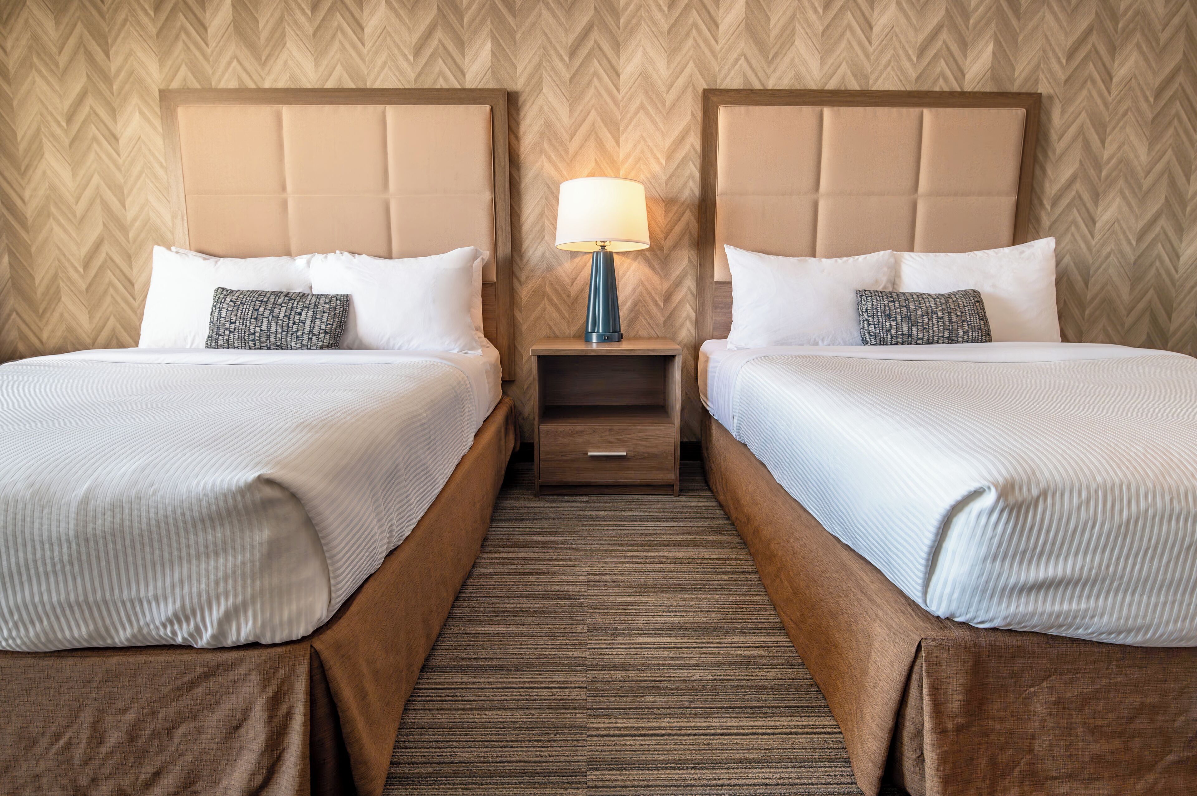 A hotel room with two beds, a nightstand with a lamp, and a herringbone-patterned wall.