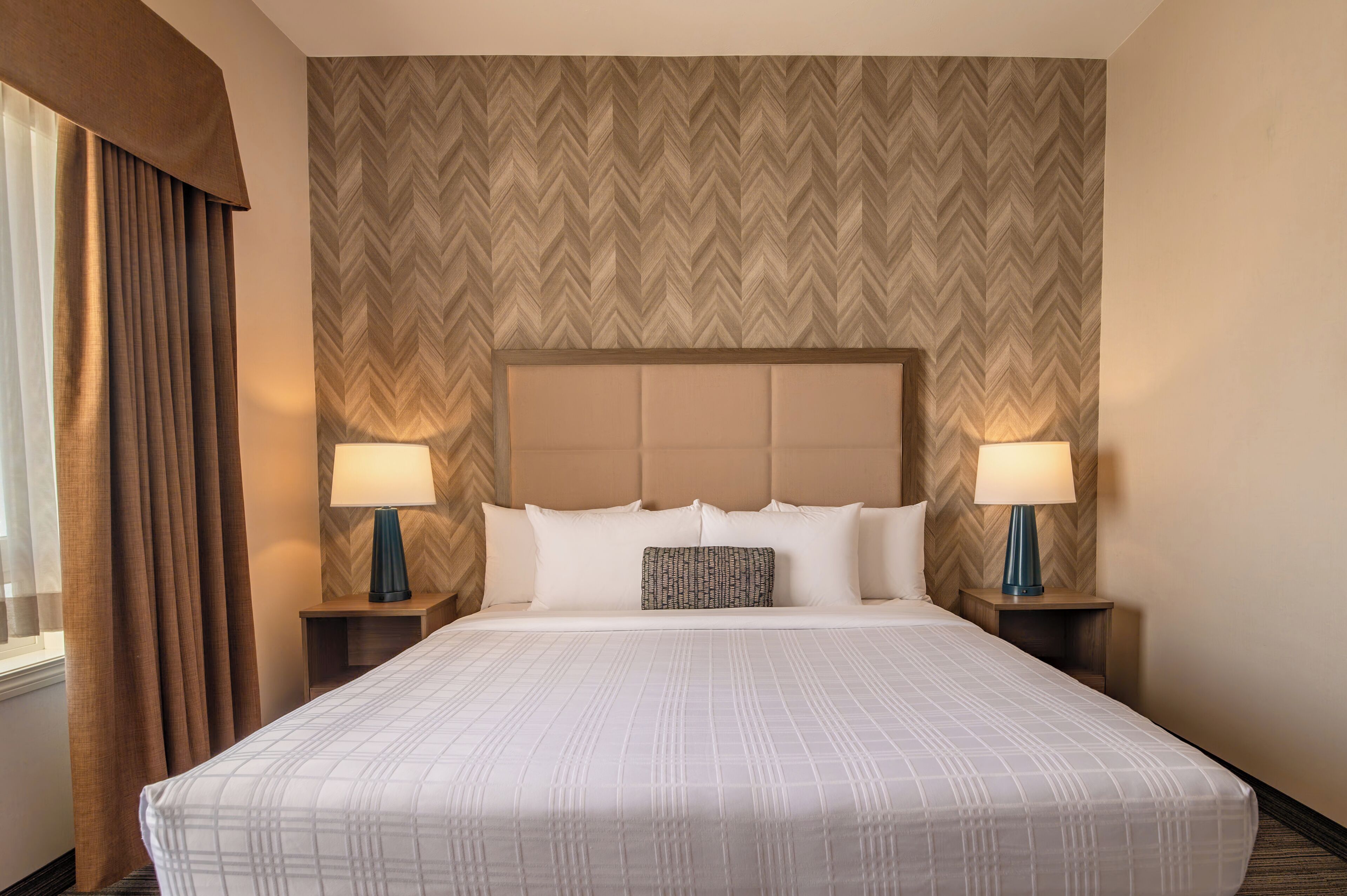 Una habitación de hotel con una cama grande, dos mesitas de noche con lámparas, un papel tapiz estampado y una ventana con cortinas.