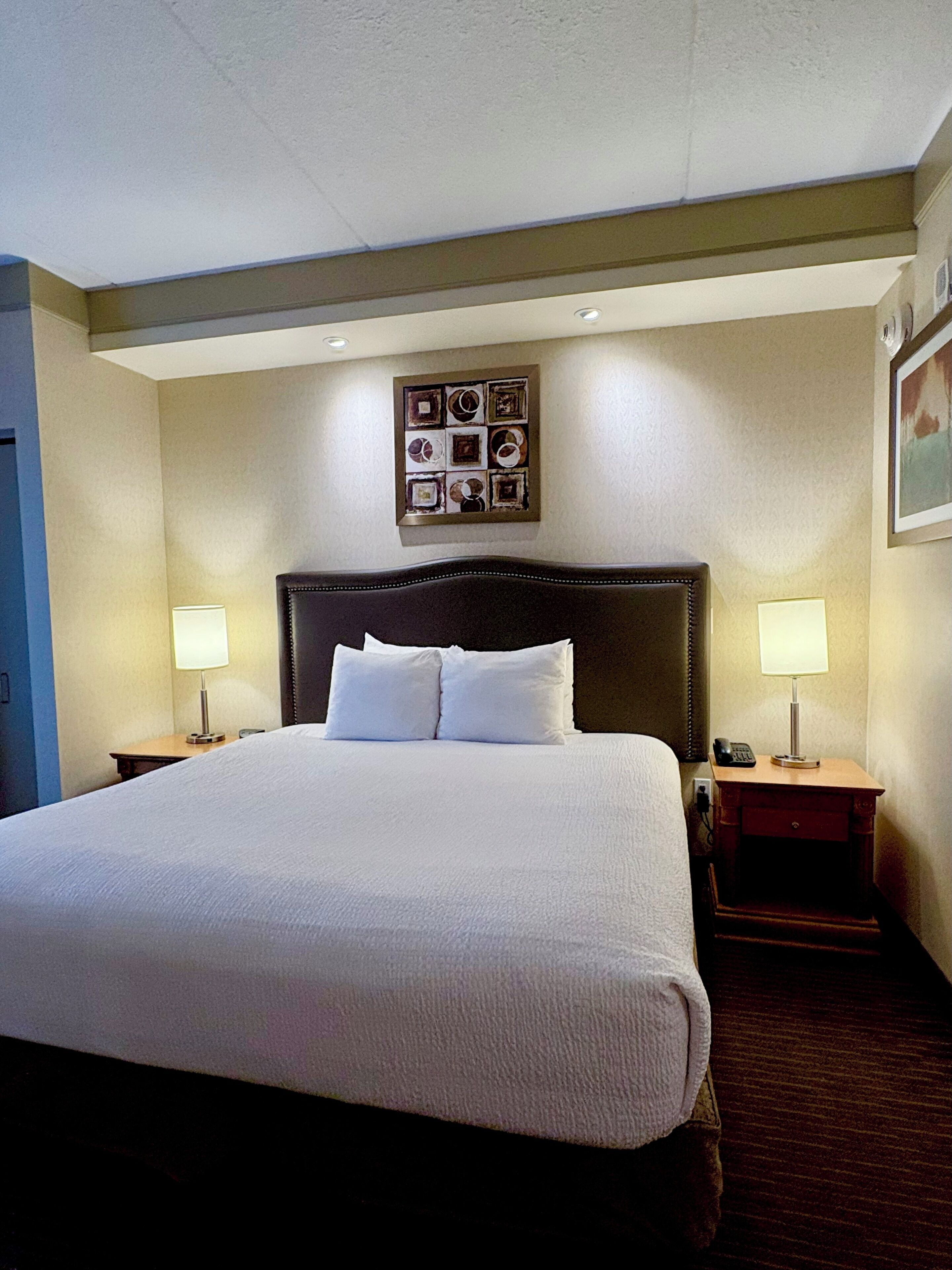 A hotel room with a large bed, two bedside lamps, a nightstand with a phone, and a framed picture on the wall.