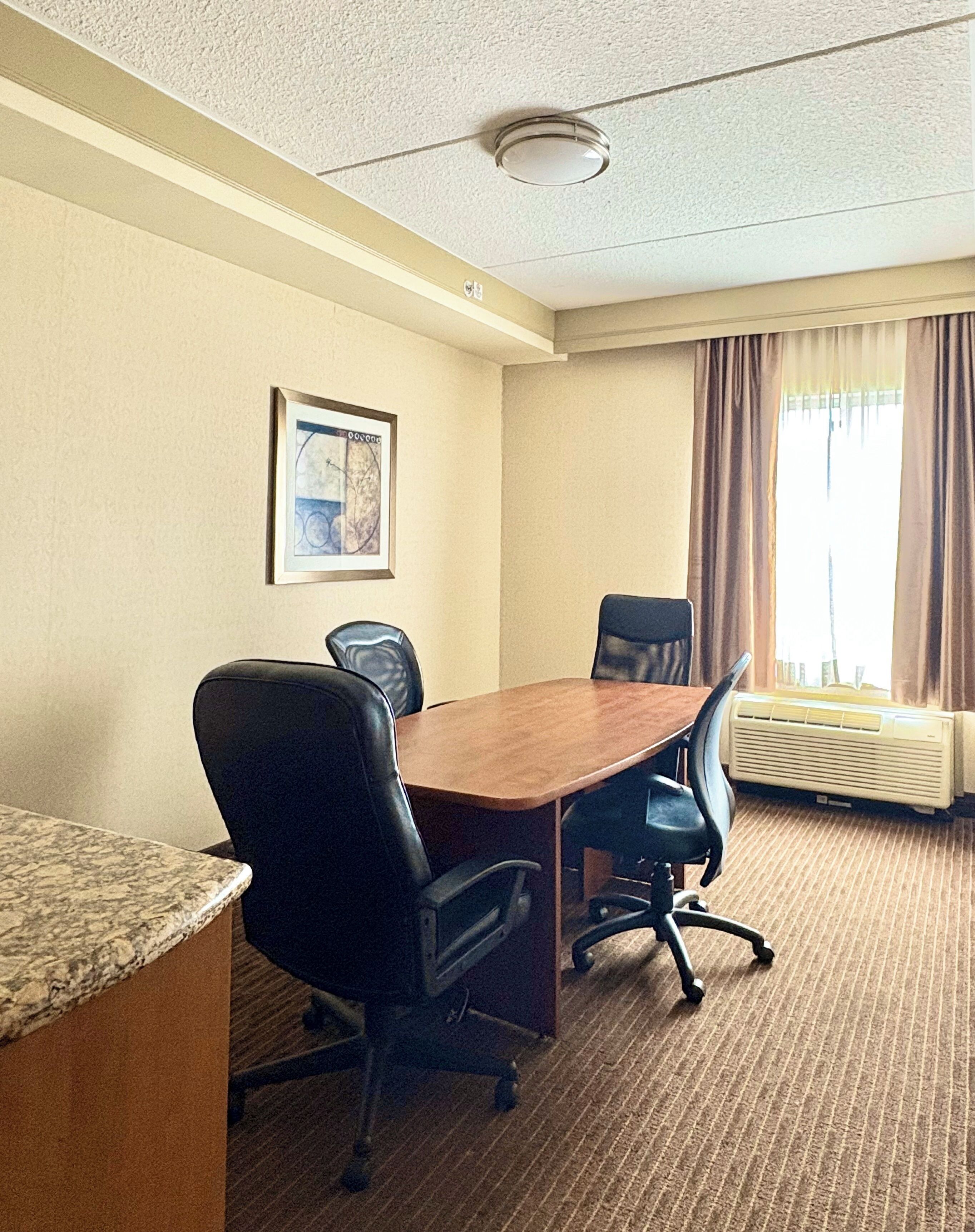 A conference room with a wooden table, four black office chairs, a framed picture on the wall, and a window with curtains.