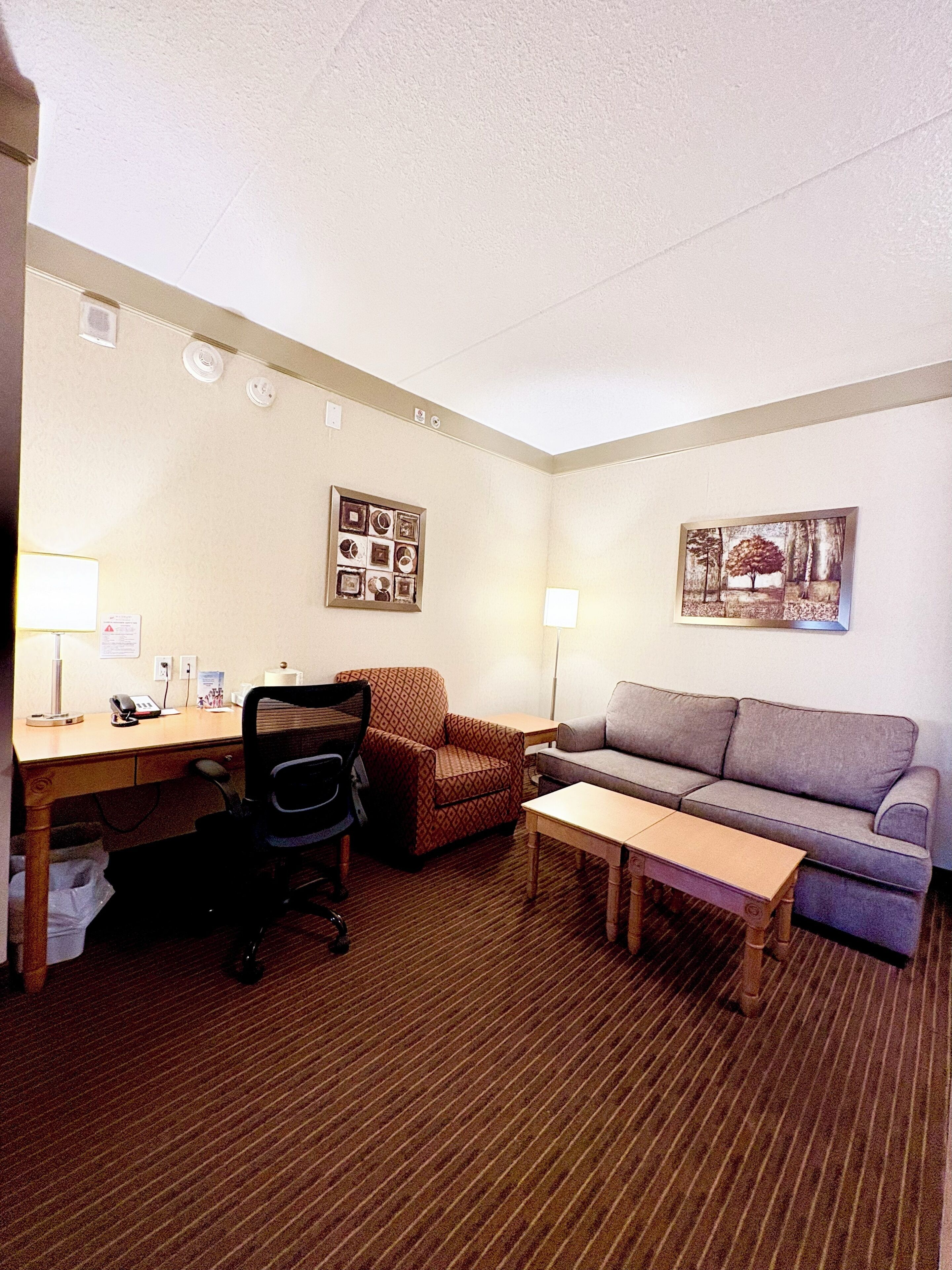 A hotel room with a desk, office chair, sofa, and a small table. There are two wall-mounted lamps, a framed picture, and a bulletin board with various items pinned to it.