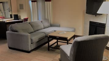 Un salon moderne avec un canapé d’angle gris, une petite table basse en bois et un fauteuil gris. Un téléviseur à écran plat est fixé au mur, et il y a une cheminée.
