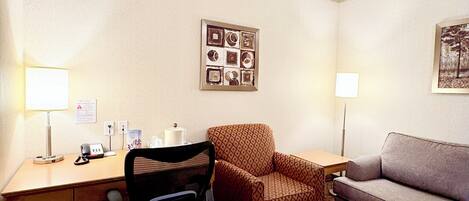 A hotel room with a desk, a chair, a sofa, a lamp, and a picture on the wall.