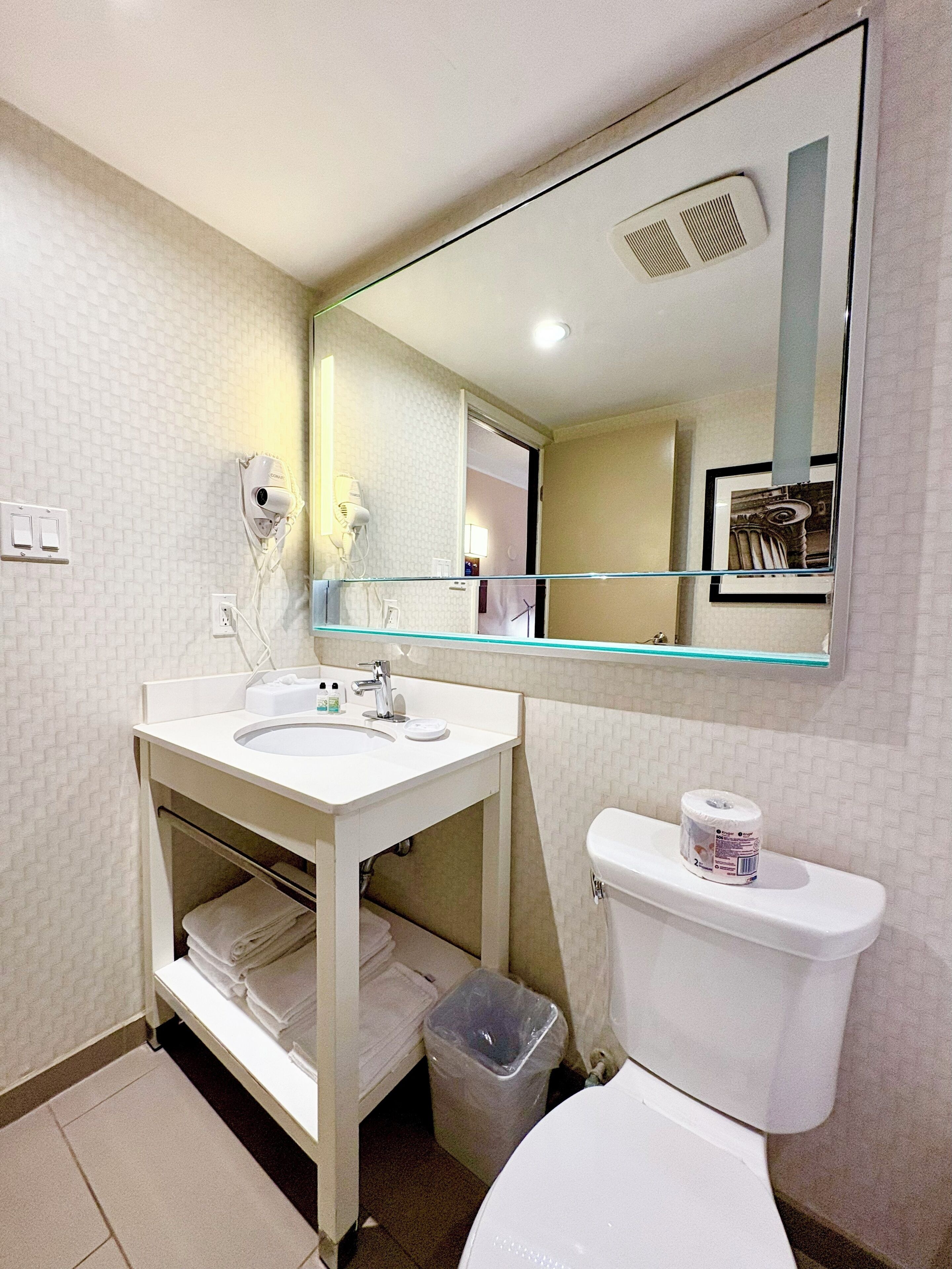 A bathroom with a white sink, mirror, and toilet. There is a trash can and a towel rack.
