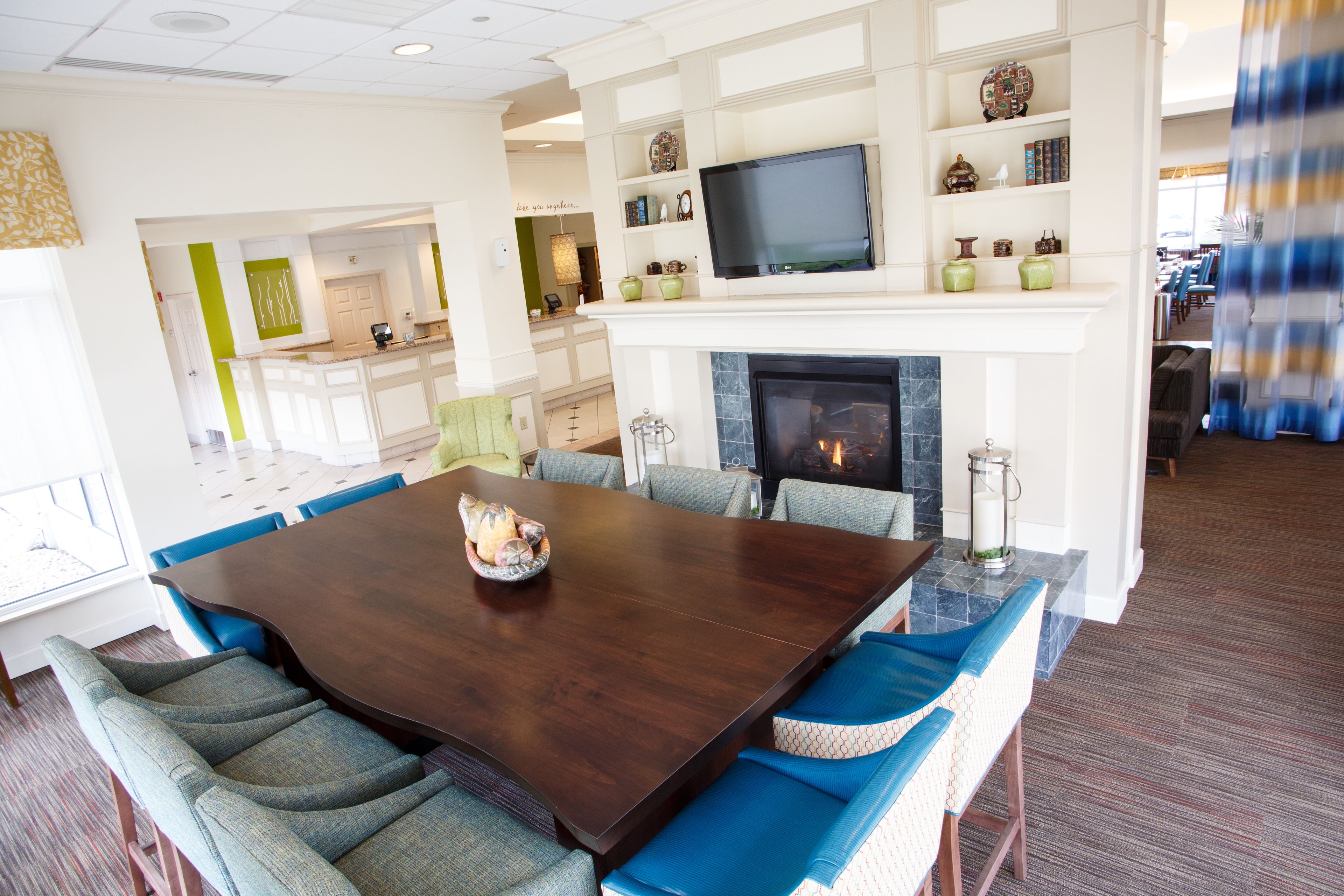 A dining area with a large wooden table and blue upholstered chairs, a fireplace, and a television mounted above it.