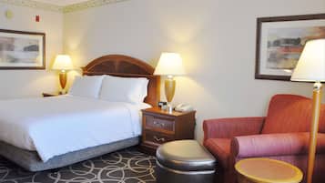 Una habitación de hotel con una cama, dos mesitas de noche con lámparas, una silla, una mesita redonda y un cubo de basura.