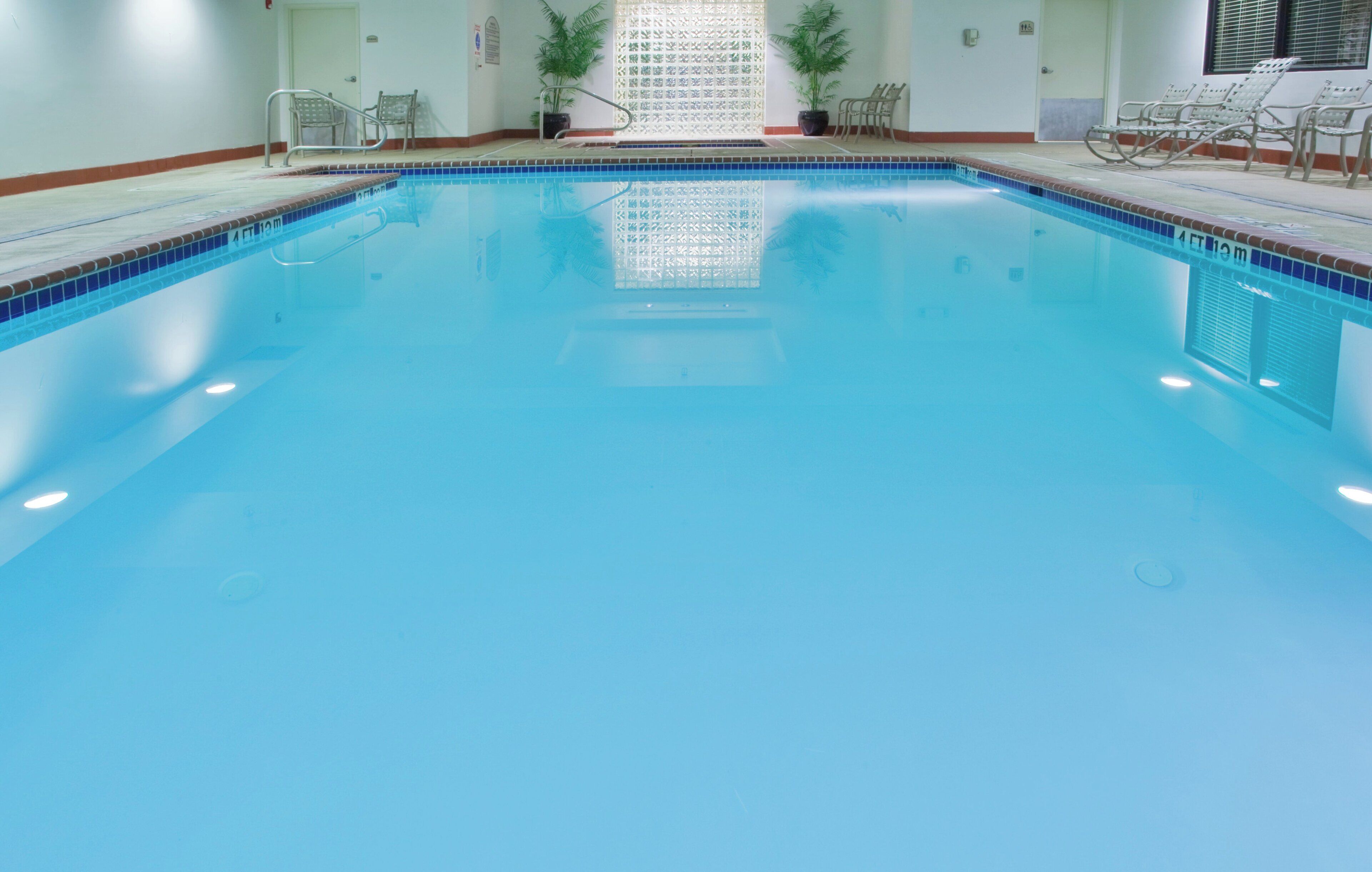 Uma piscina interior translúcida, com borda azulejada, escada de metal e cadeiras ao redor.