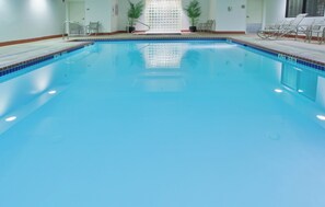 Una piscina interior luminosa con borde de azulejos, una escalera metálica y sillas alrededor del perímetro.