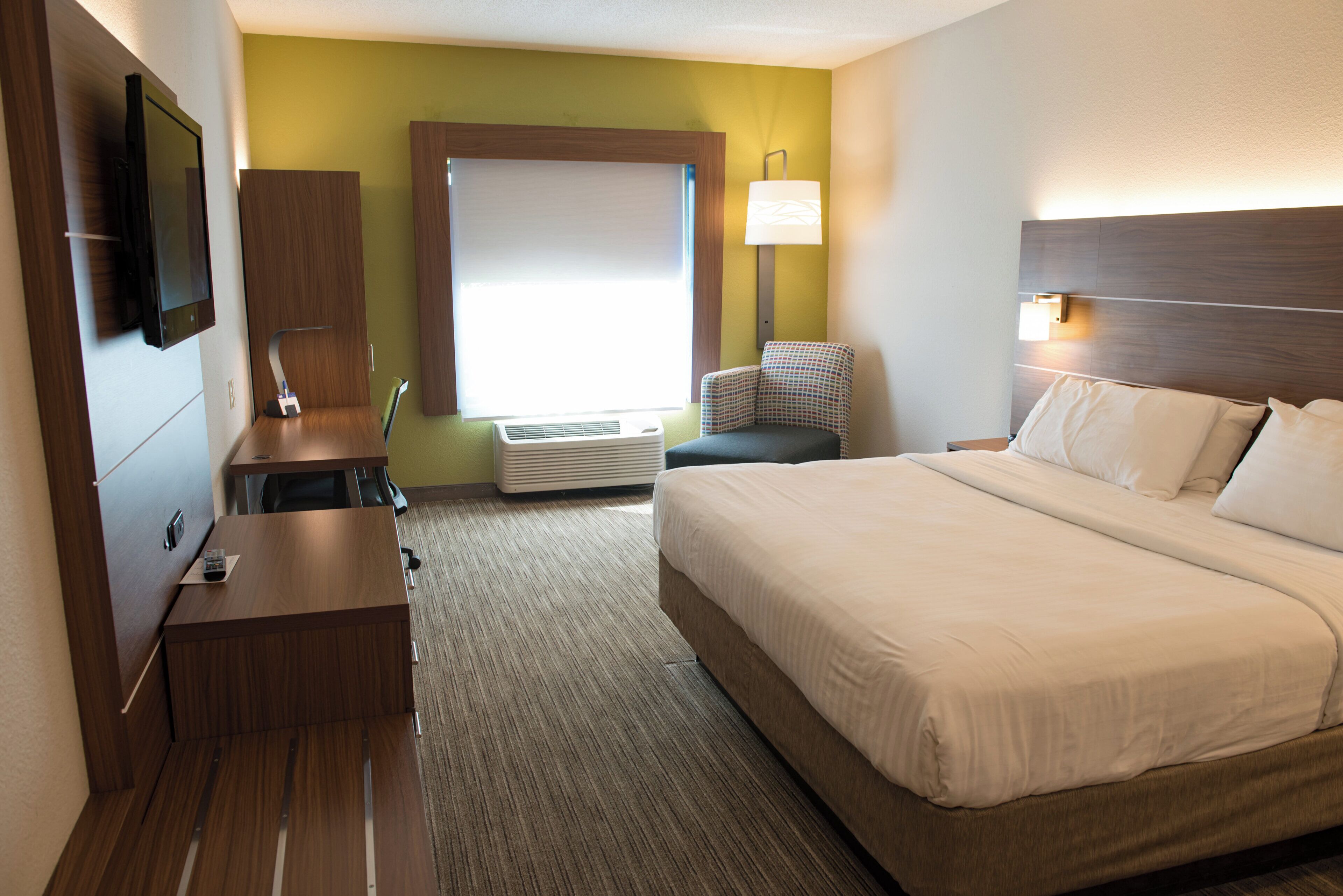 Habitación de hotel con cama, escritorio, silla, televisor, aire acondicionado y ventana con persianas.