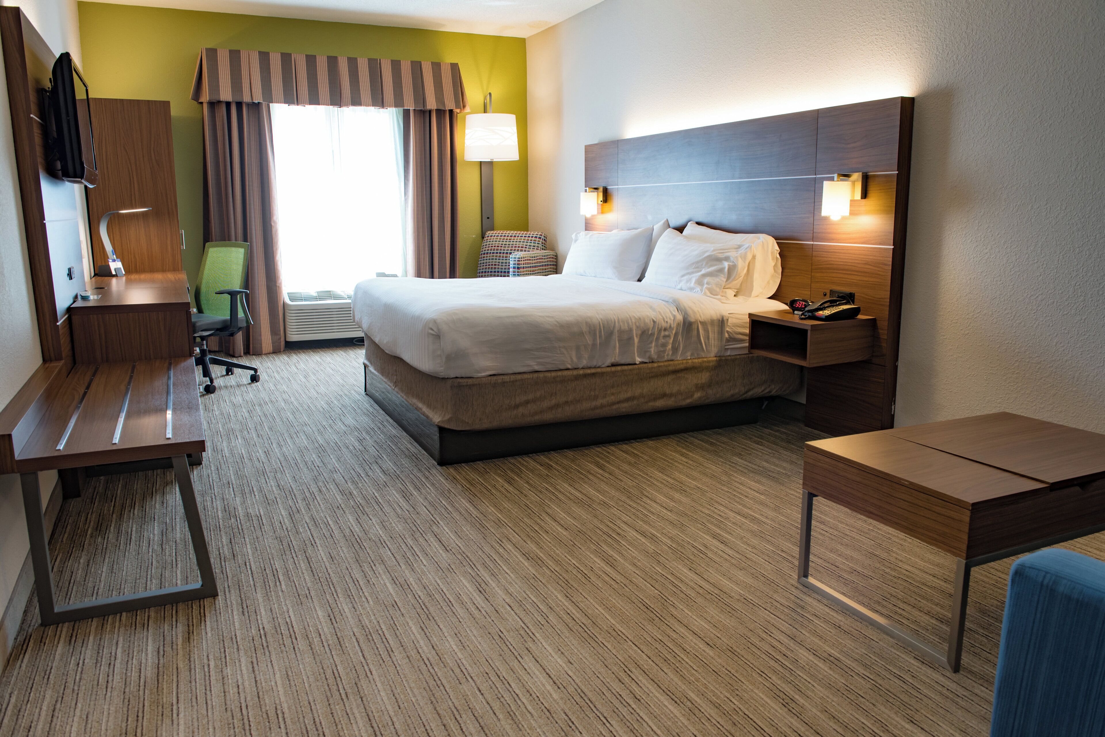 A hotel room with a bed, bedside tables, a desk, a chair, a lamp, and a window with curtains.