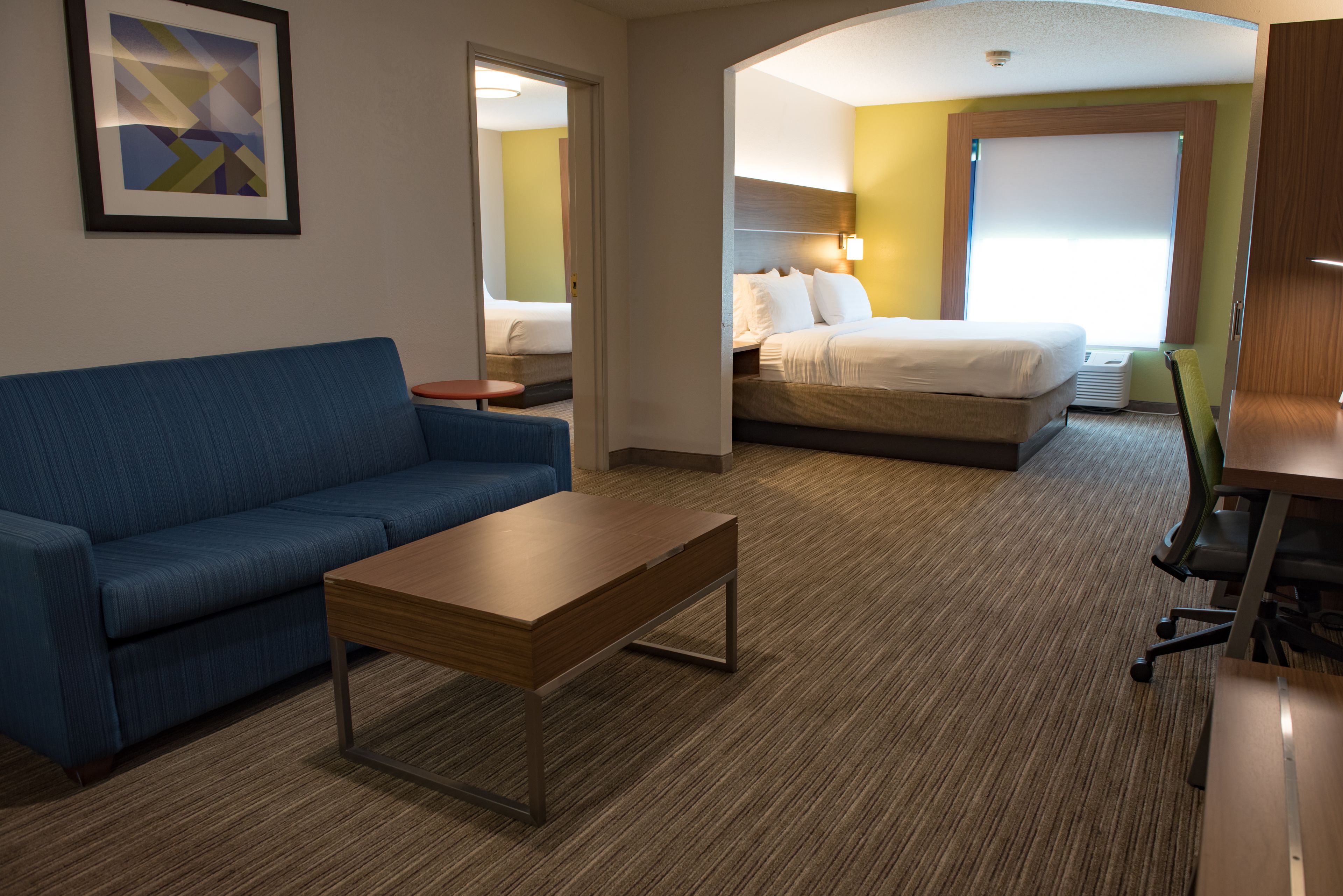 A hotel room with a blue sofa, a wooden coffee table, a bed with white linens, and a large window.