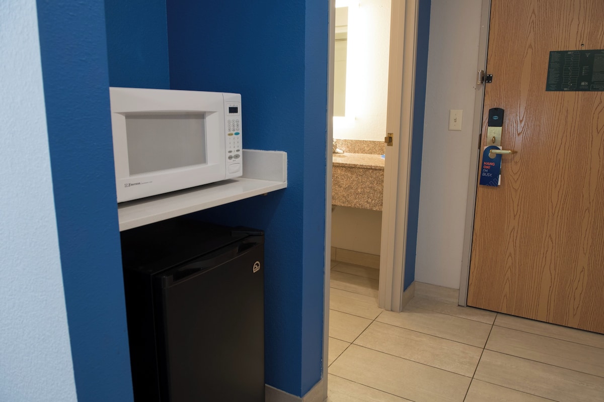 A compact kitchenette with a microwave and a small refrigerator, a wooden door with a lock, and a bathroom visible through an open door.