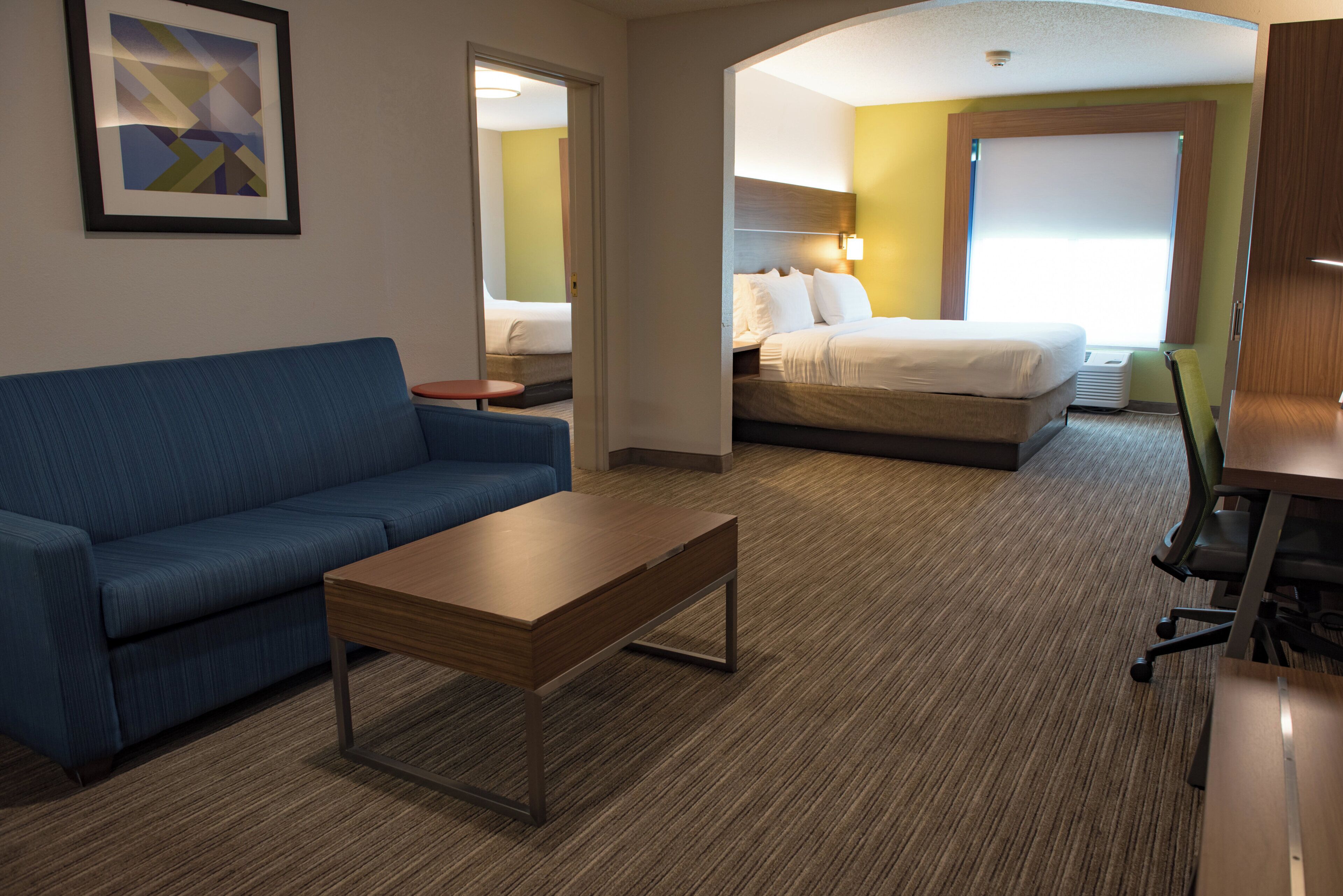 A hotel room with a blue sofa, a wooden coffee table, a bed with white bedding, and a desk with a chair.