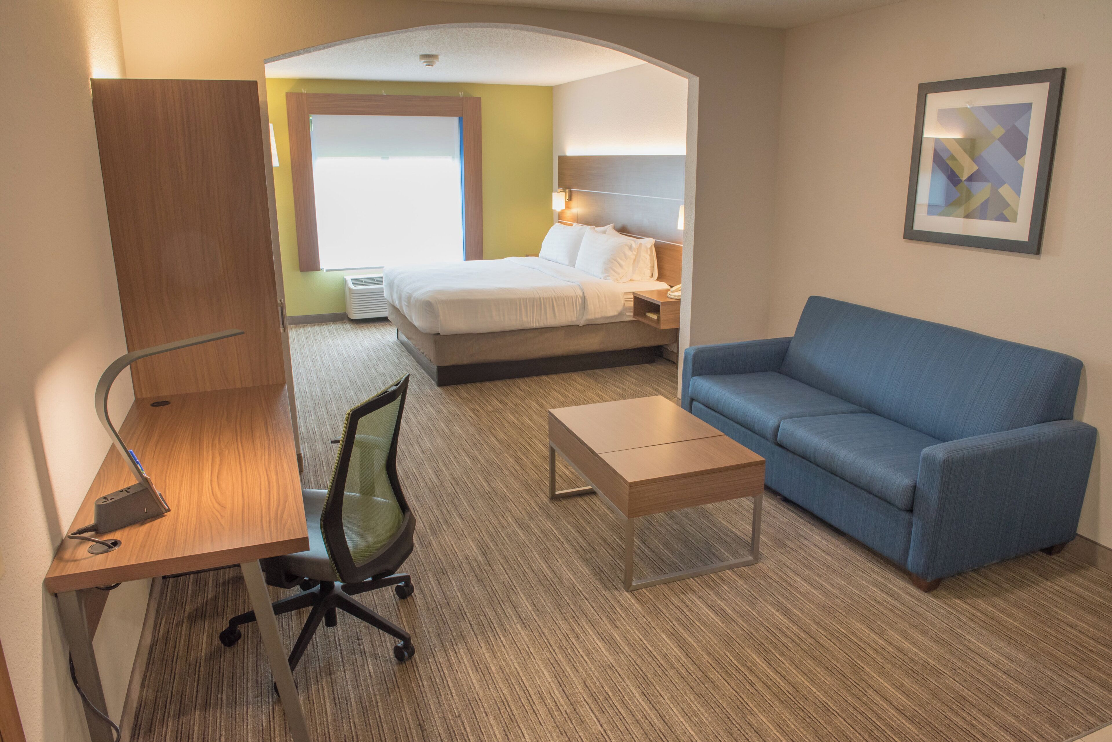 A hotel room with a bed, a blue sofa, a desk with a chair, and a small table.