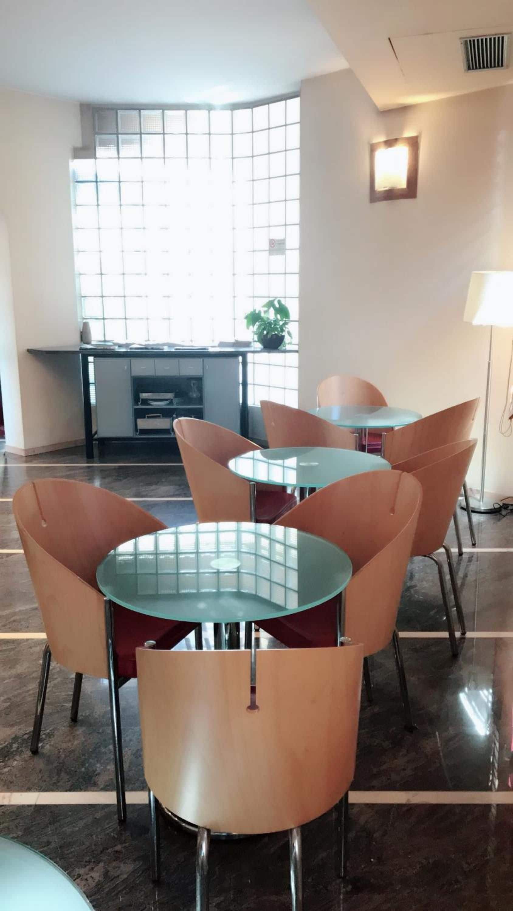 A modern dining area with glass-top tables and orange chairs, a kitchen in the background, and a large window providing natural light.