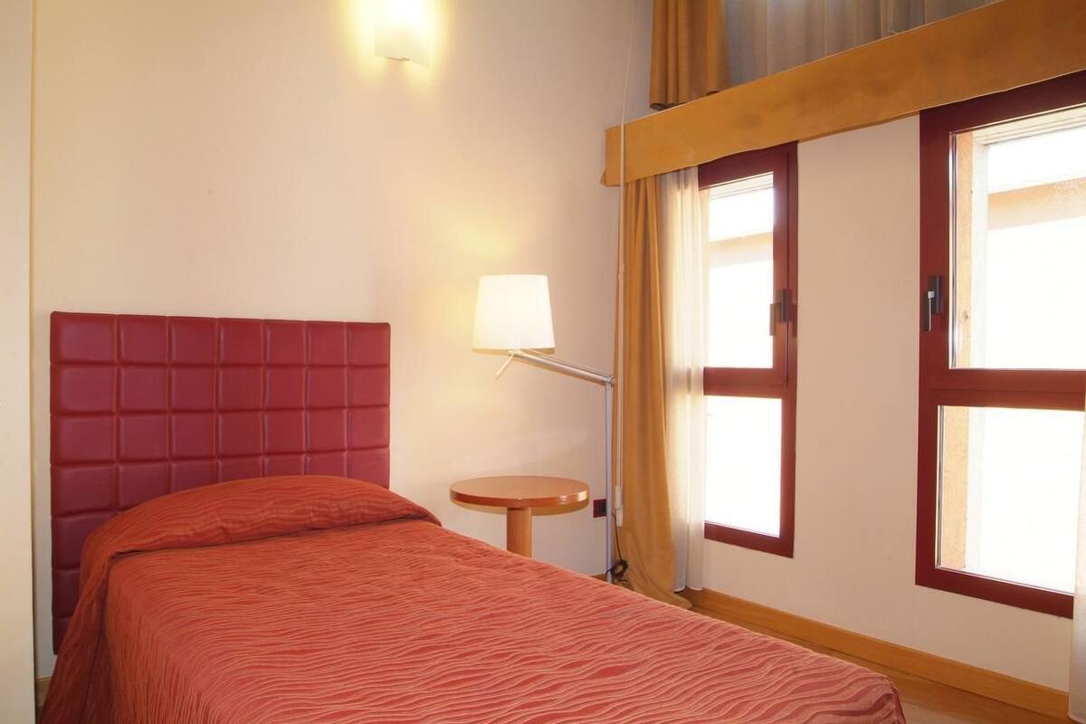 A hotel room with a red headboard, a small round table, a lamp, and a window with curtains.