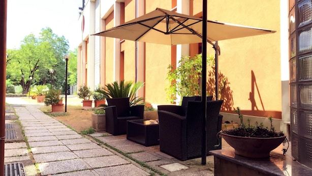 Un área de estar al aire libre con mobiliario de mimbre y un gran paraguas, rodeada de plantas en macetas y un edificio con columnas.