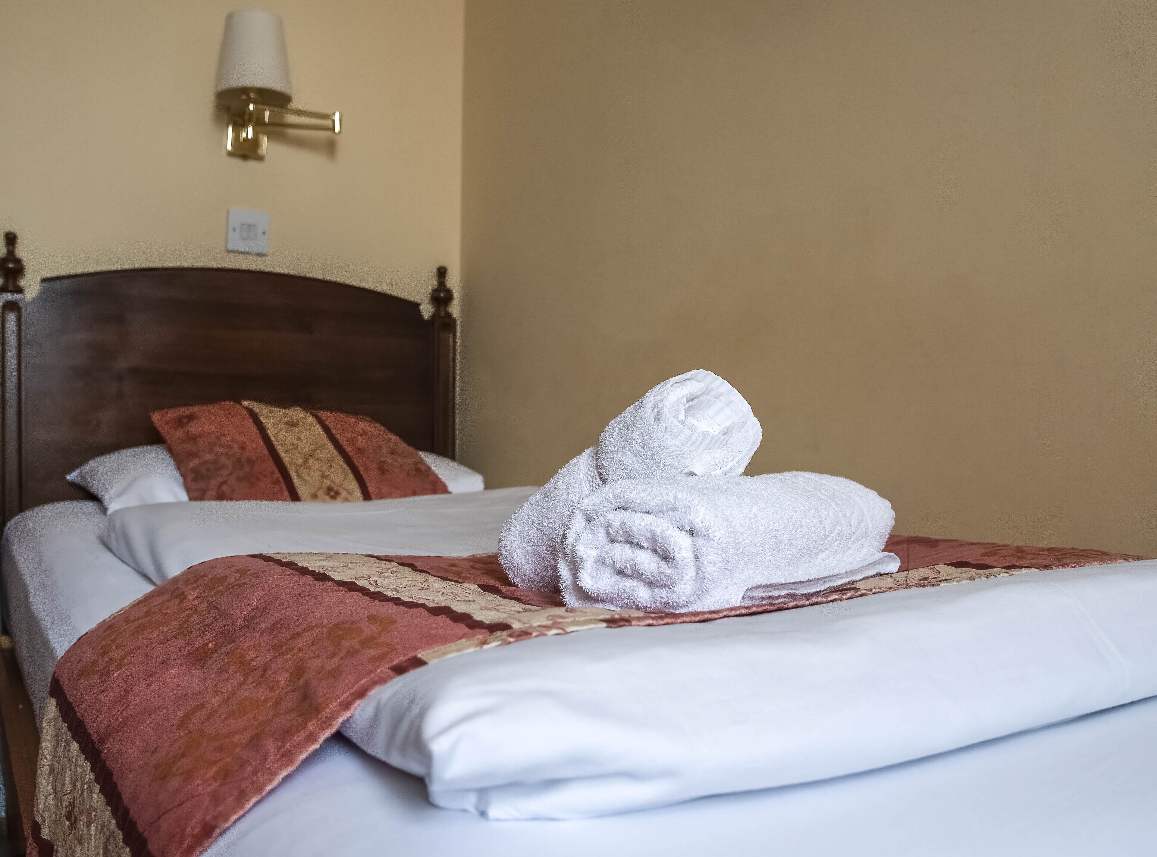 A neatly made bed with white linens, a red and beige patterned throw blanket, and two white towels folded on top.
