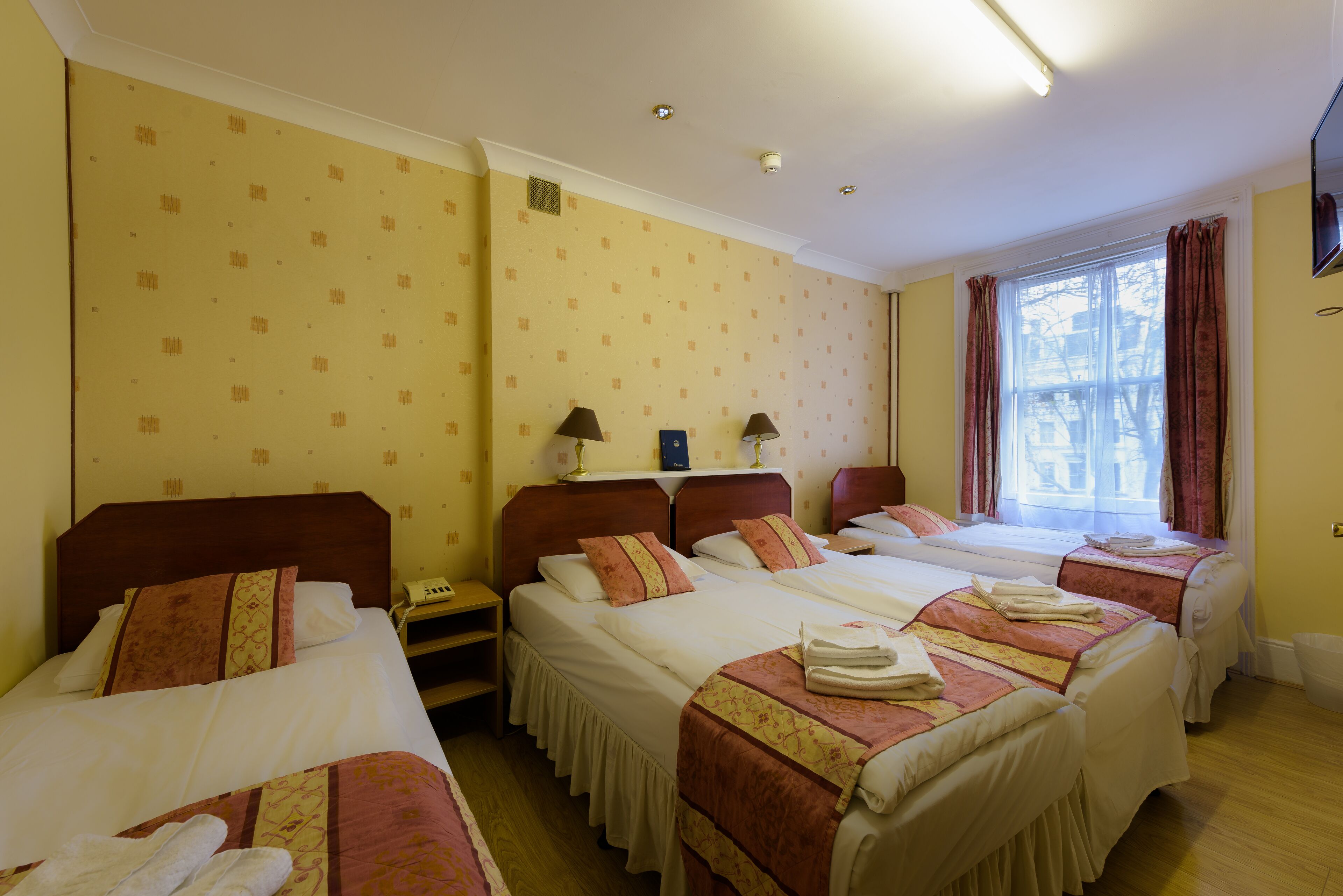 A hotel room with three beds, each with a different style of bedding and headboards. There is a window with curtains and a view of the outside.