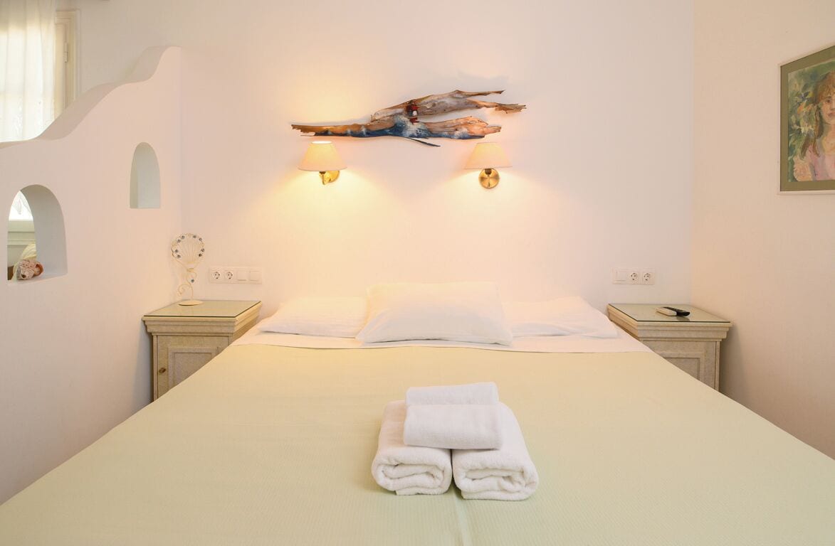 A neatly made bed with white linens and a folded towel, two bedside tables with lamps, a wall-mounted artwork, and a small mirror.