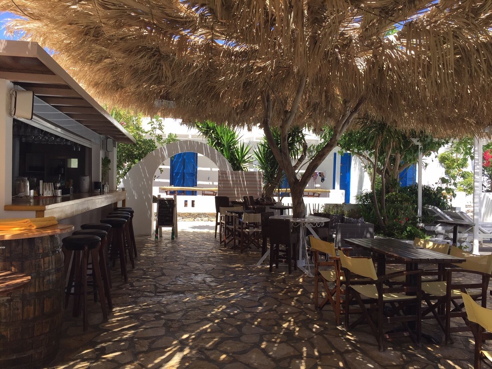 An outdoor café with stone flooring, wooden tables and chairs, a bar area, and a thatched roof.