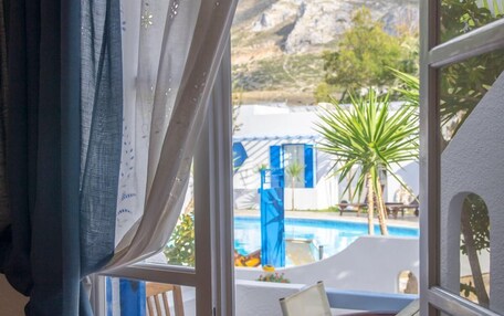 A room with a view of a swimming pool, a palm tree, and a mountainous landscape.