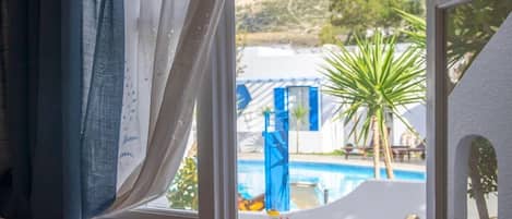 A room with a view of a swimming pool, a palm tree, and a mountainous landscape.