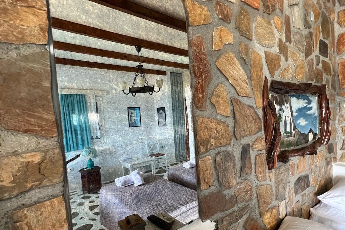 Ein Zimmer mit steinernen Wänden, ausgestattet mit einem Bett, einem Spiegel, einer Kristallleuchtre und einem kleinen Tisch mit Lampe und Fernbedienungen.