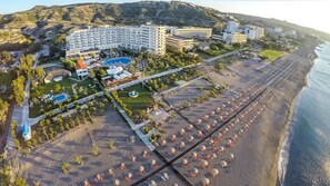 Luftaufnahme eines Küstenhotels mit mehreren Gebäuden, einem Swimmingpool und einem Strand mit Sonnenschirmen.