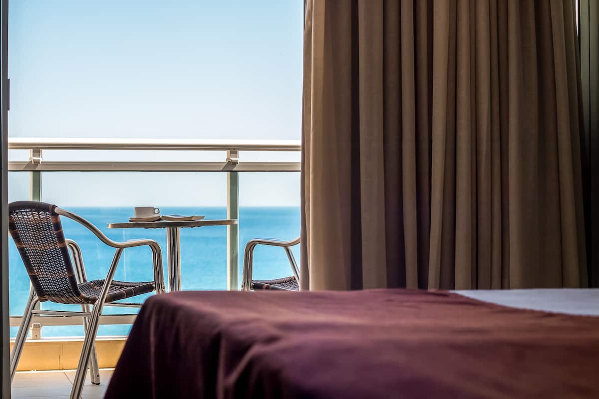 Une chambre d’hôtel avec un balcon, un lit et deux chaises donnant sur l’océan.