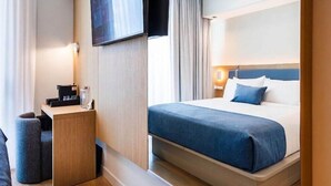 A hotel room with a bed, a desk with a lamp, a TV mounted on the wall, and a window with curtains. - Sanctuary South Beach (MIAMI BEACH)