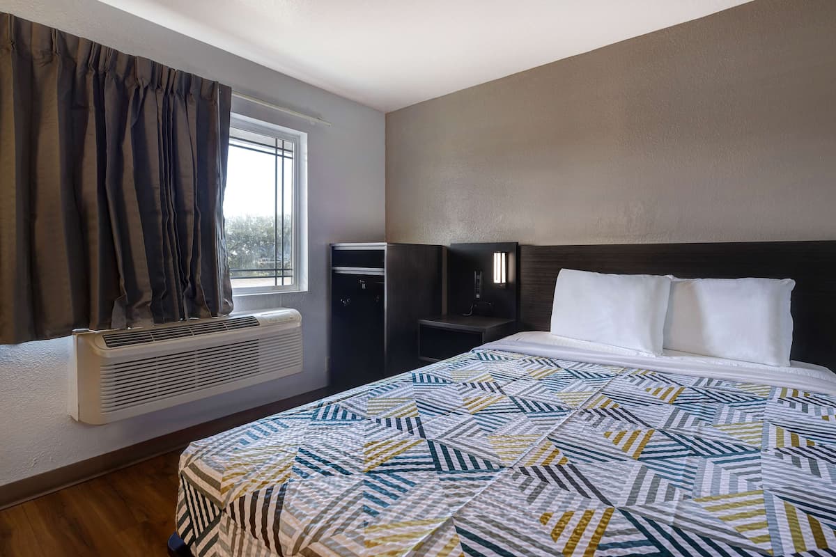 A hotel room with a large bed, a window with curtains, a heater, a small table, and a dark-colored wardrobe.